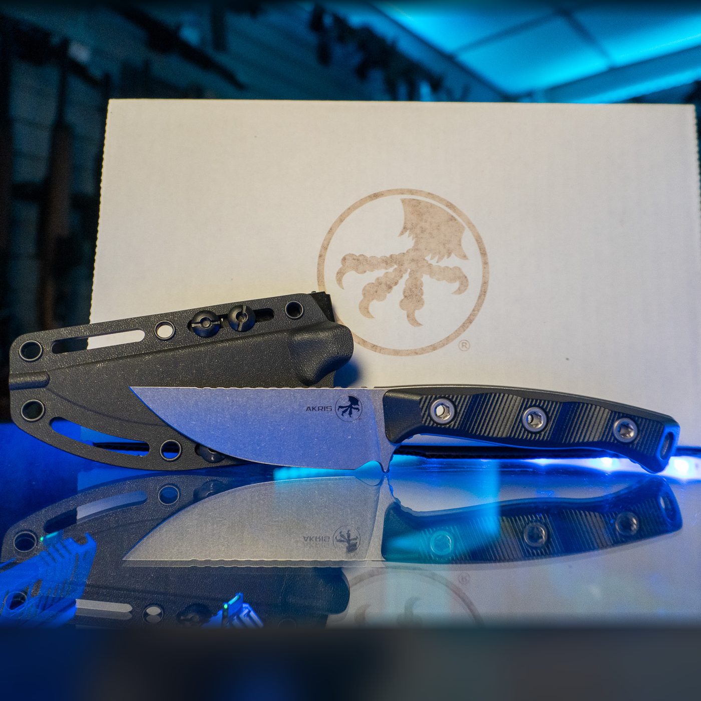 Microtech Akris fixed blade knife with stonewash drop point blade and black G-10 handle, shown with its black Kydex sheath and Microtech box