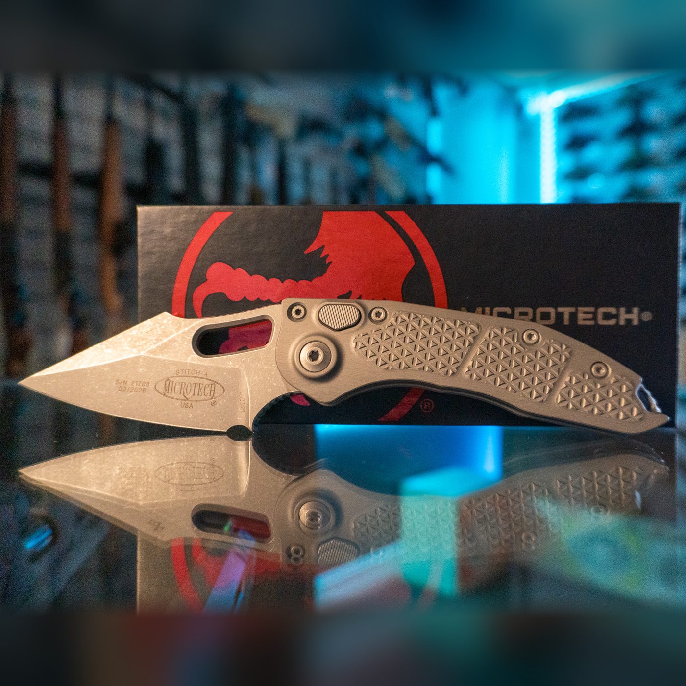 Microtech Stitch-A automatic knife with Apocalyptic finish spear point blade and textured natural clear aluminum handle, in front of its Microtech box