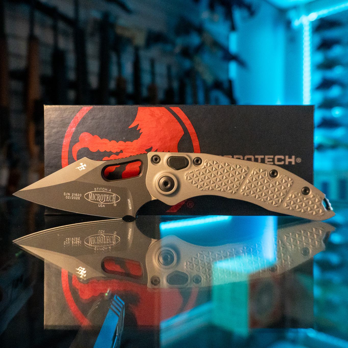 Microtech Stitch-A automatic knife with black spear point blade and textured natural clear aluminum handle, standing in front of its Microtech box