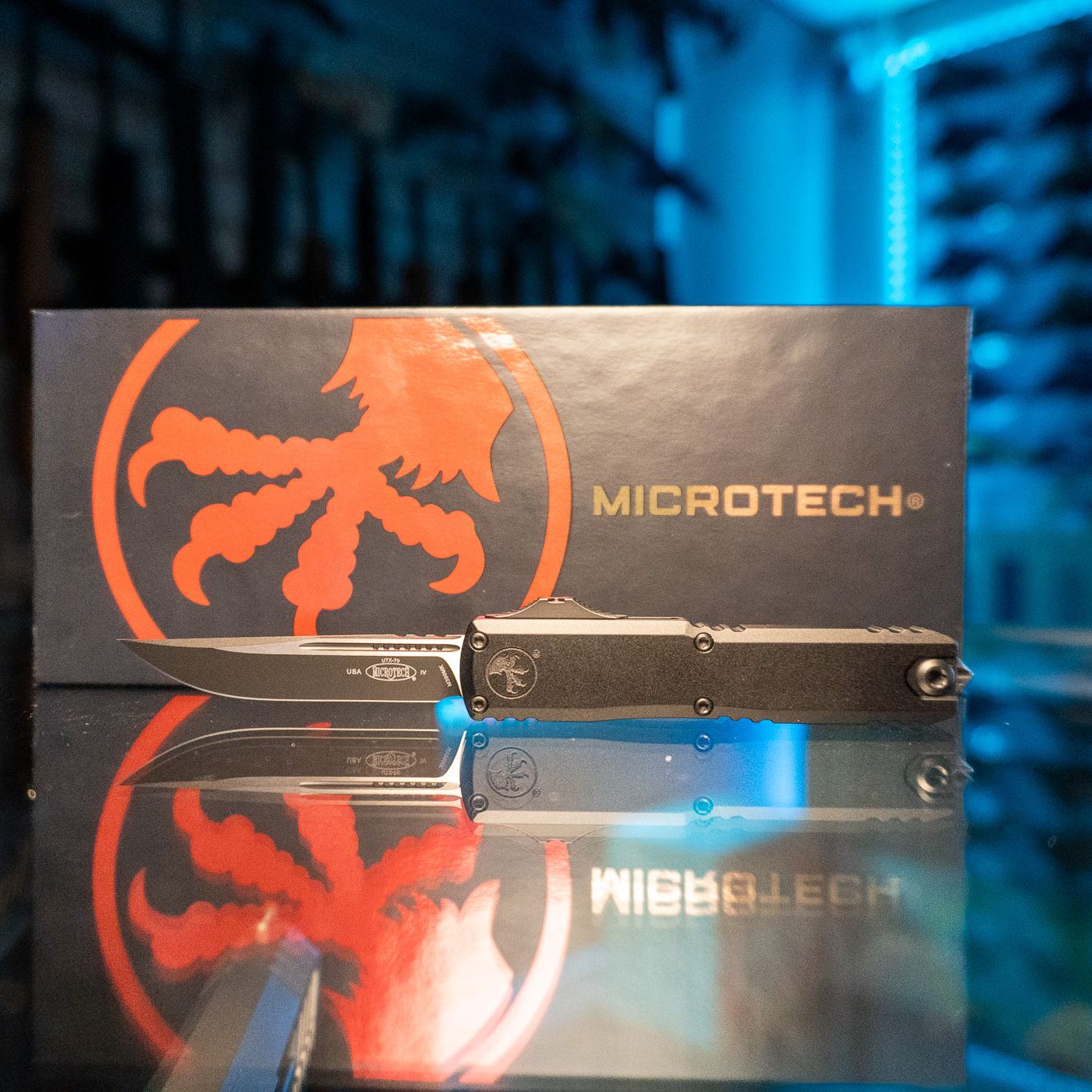 Microtech UTX-70 Gen IV OTF knife with black single-edge spear point blade deployed and black aluminum handle, in front of its Microtech box