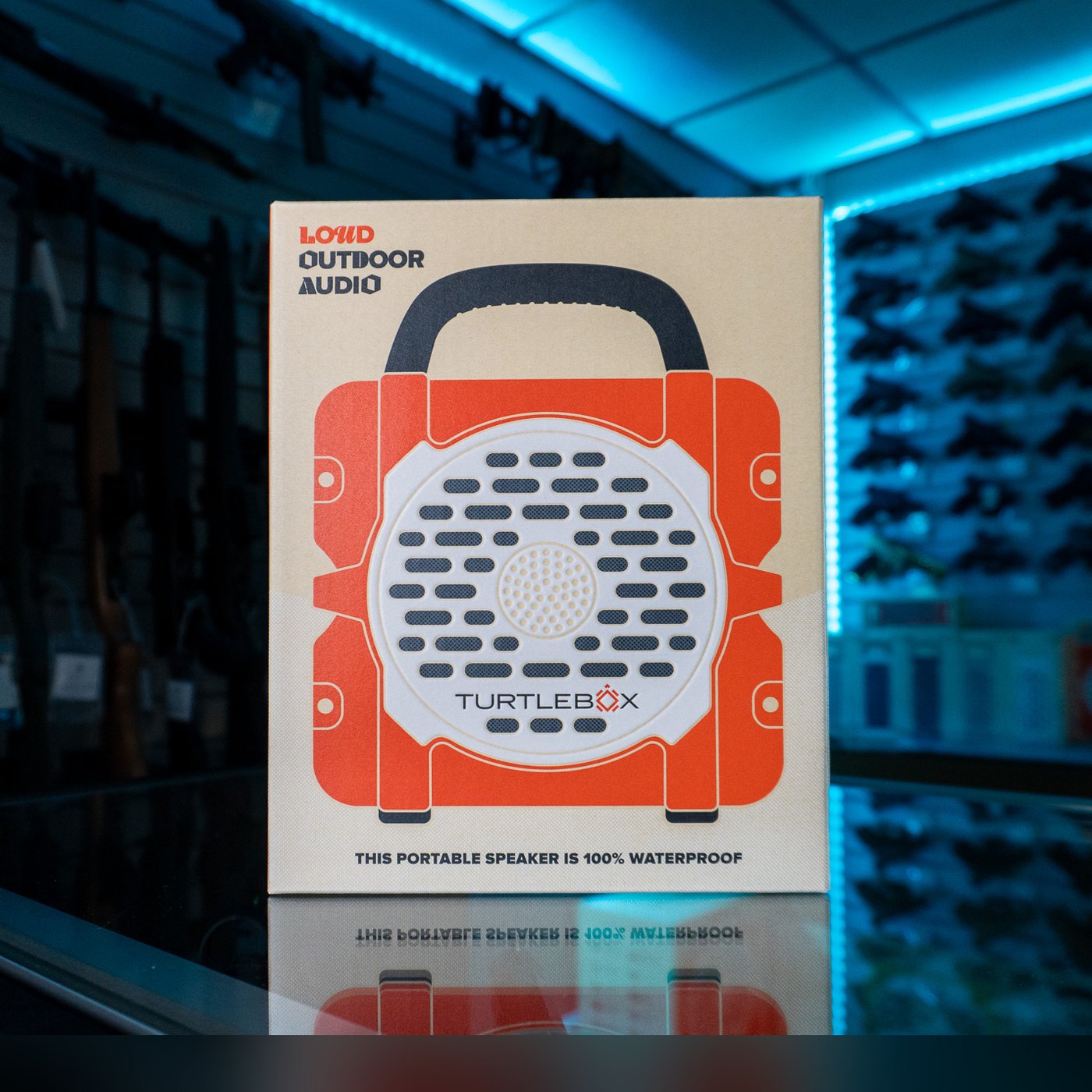 Turtlebox Cub speaker retail box with an orange and white speaker illustration and black handle, printed "Loud Outdoor Audio", on a glass display counter