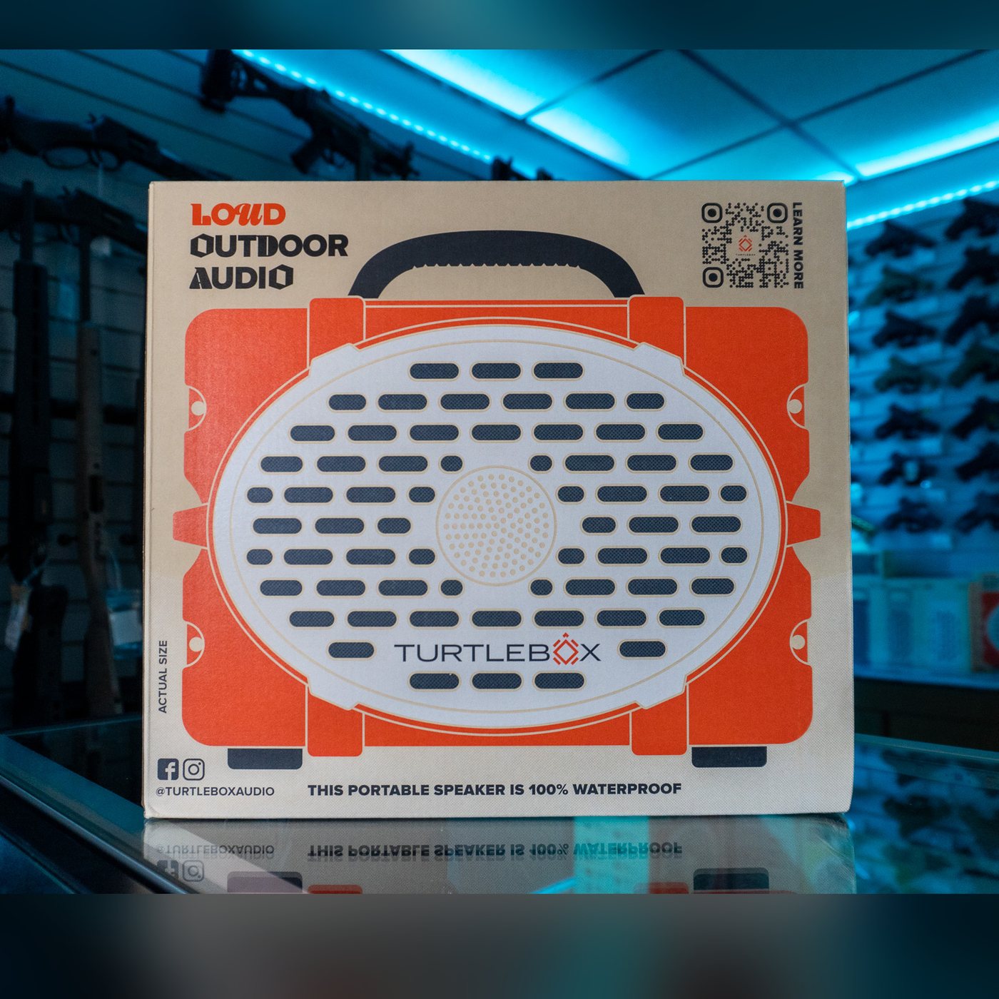 Turtlebox Original Gen 3 speaker retail box with an orange and white speaker illustration and black handle, printed "Loud Outdoor Audio", on a glass display counter