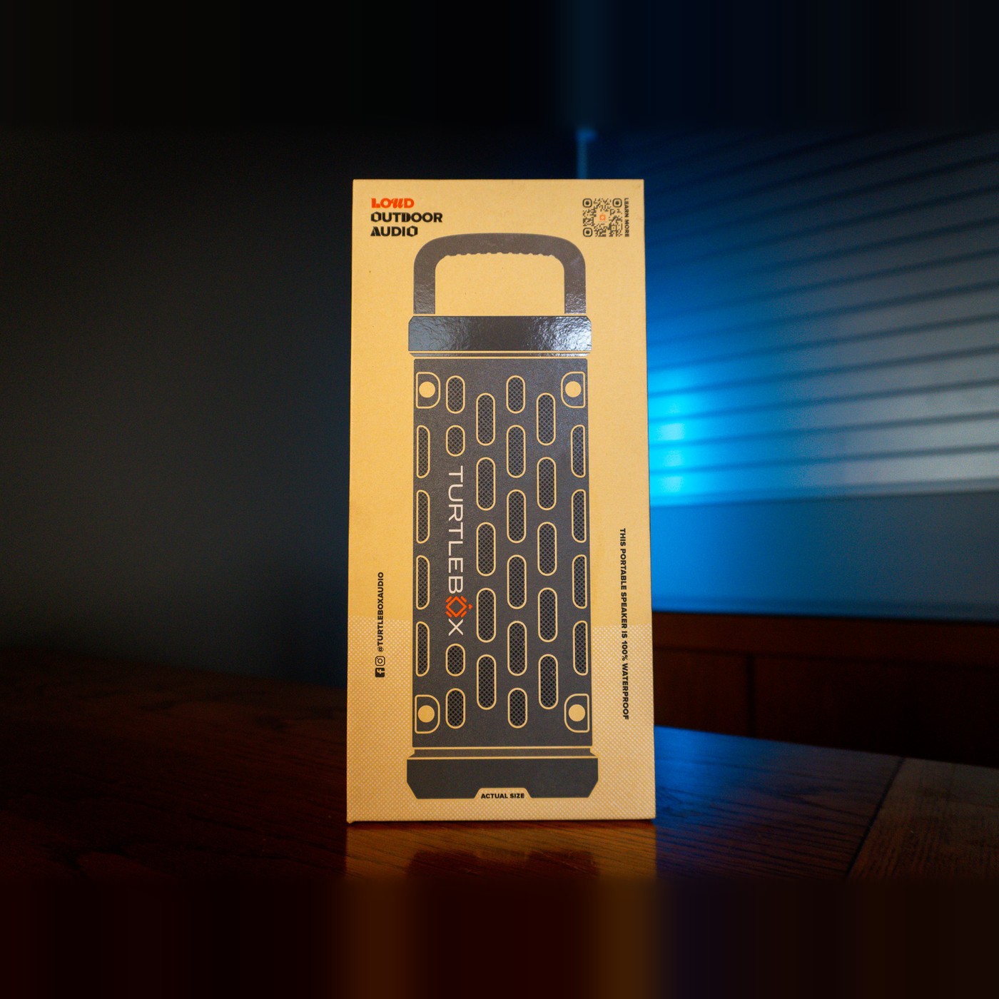 Turtlebox Ranger speaker retail box in kraft cardboard with a dark gray speaker illustration and handle, printed "Loud Outdoor Audio", on a wooden counter