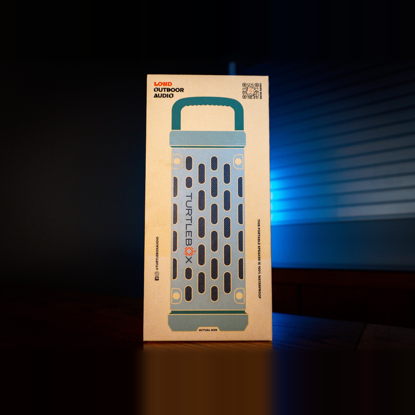 Turtlebox Ranger speaker retail box in kraft cardboard with a light blue speaker illustration and teal handle, printed "Loud Outdoor Audio", on a wooden counter